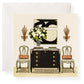 A formal room features a black sideboard, two chairs with red cushions, a vase of white flowers, Karen Adams Dolce Vita Gift Enclosures displayed elegantly, a monochrome artwork, and two wall sconces.