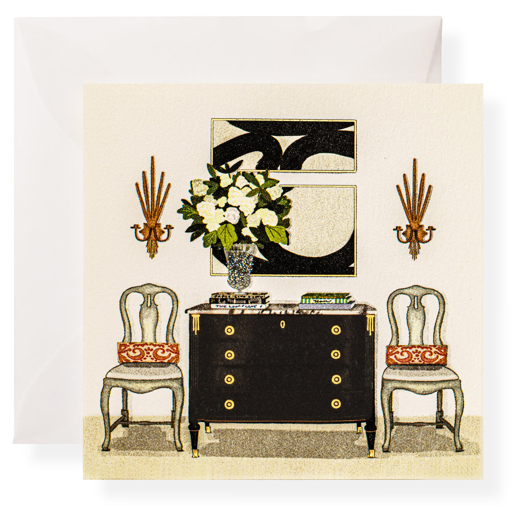 A formal room features a black sideboard, two chairs with red cushions, a vase of white flowers, Karen Adams Dolce Vita Gift Enclosures displayed elegantly, a monochrome artwork, and two wall sconces.