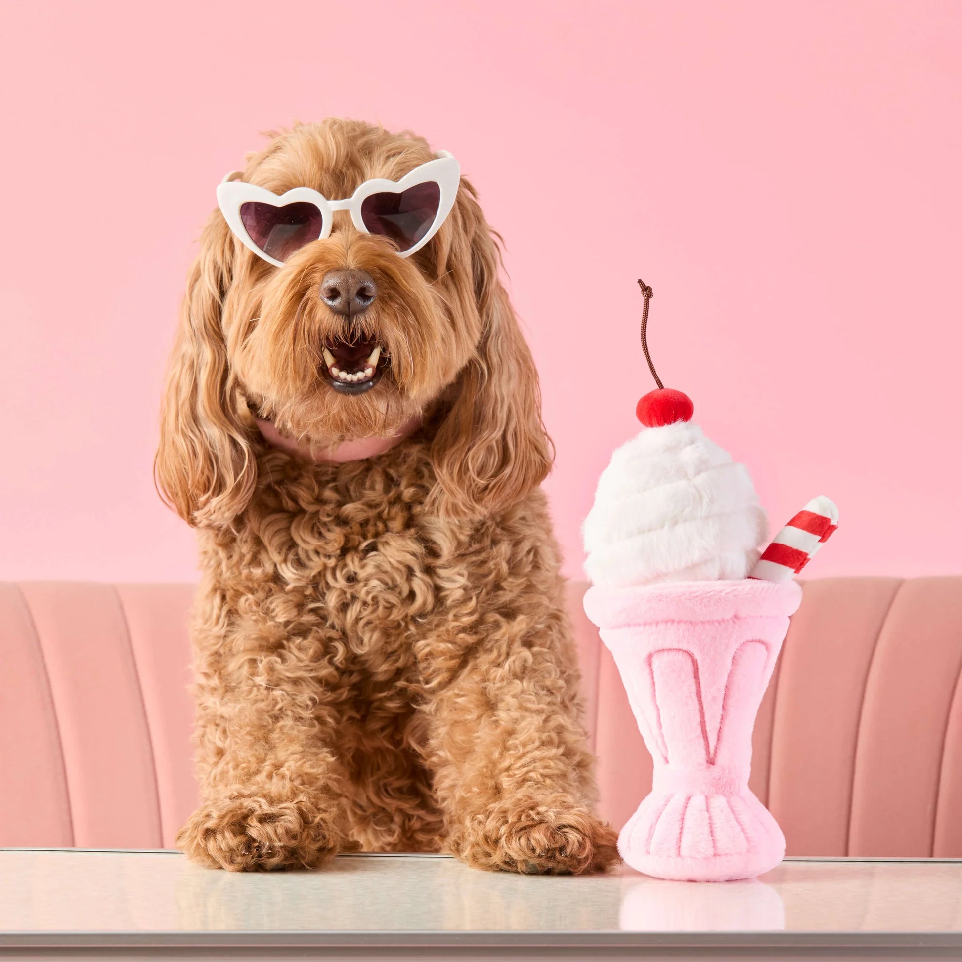 A curly-haired dog in heart-shaped sunglasses sits beside Foggy Dog's Strawberry Milkshake Snuffle Dog Toy, ready for playtime on a pink background.