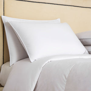 Two Frette Cortina Medium Density Pillow Inserts in white, crafted from luxurious cotton sateen, are stacked against a beige upholstered headboard on a neatly made bed with a white comforter.