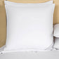 A Frette Cortina Firm Density Pillow Insert with white cotton sateen pillowcases sits on a bed with a beige headboard, accompanied by additional pillows stacked on the right side.