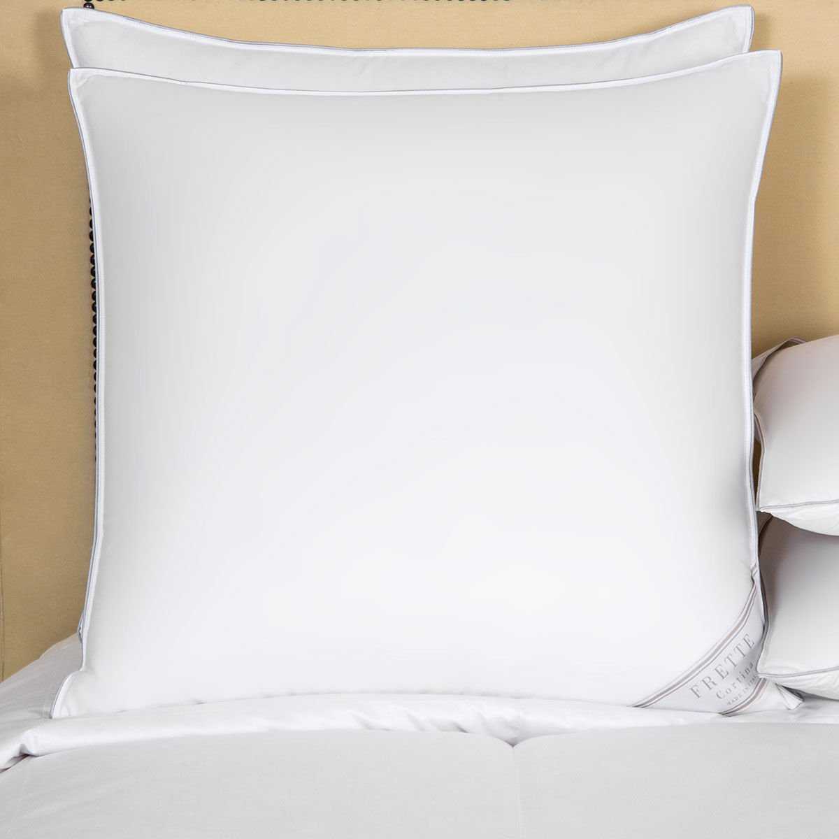 A Frette Cortina Firm Density Pillow Insert with white cotton sateen pillowcases sits on a bed with a beige headboard, accompanied by additional pillows stacked on the right side.