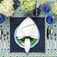 A beautifully set table features Kim Seybert's Majorelle Tablecloth Collection, showcasing handmade Indian block printing, paired with blue and white dishes, silver cutlery, blue glasses, a white napkin, and a floral centerpiece.