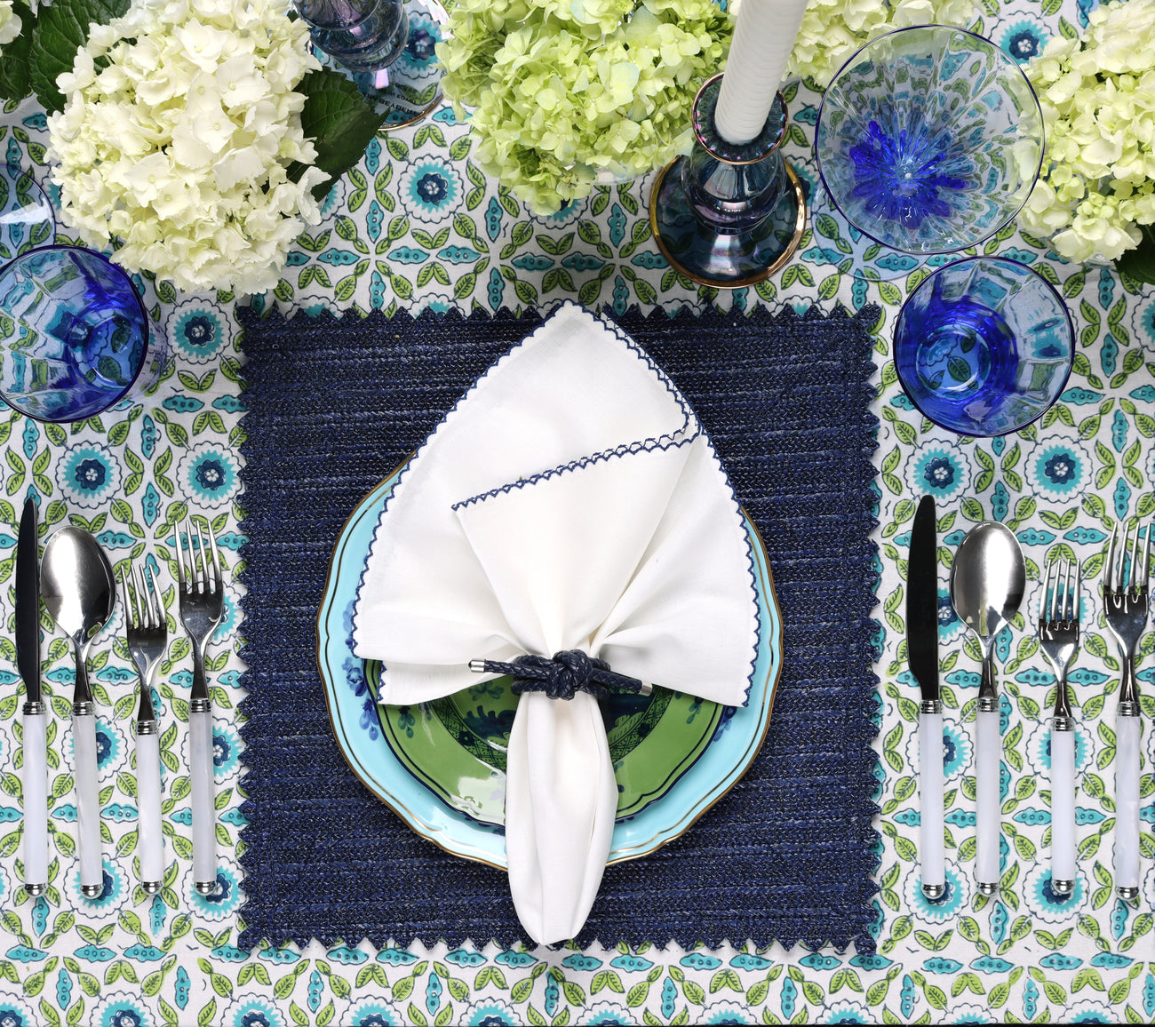 A beautifully set table features Kim Seybert's Majorelle Tablecloth Collection, showcasing handmade Indian block printing, paired with blue and white dishes, silver cutlery, blue glasses, a white napkin, and a floral centerpiece.