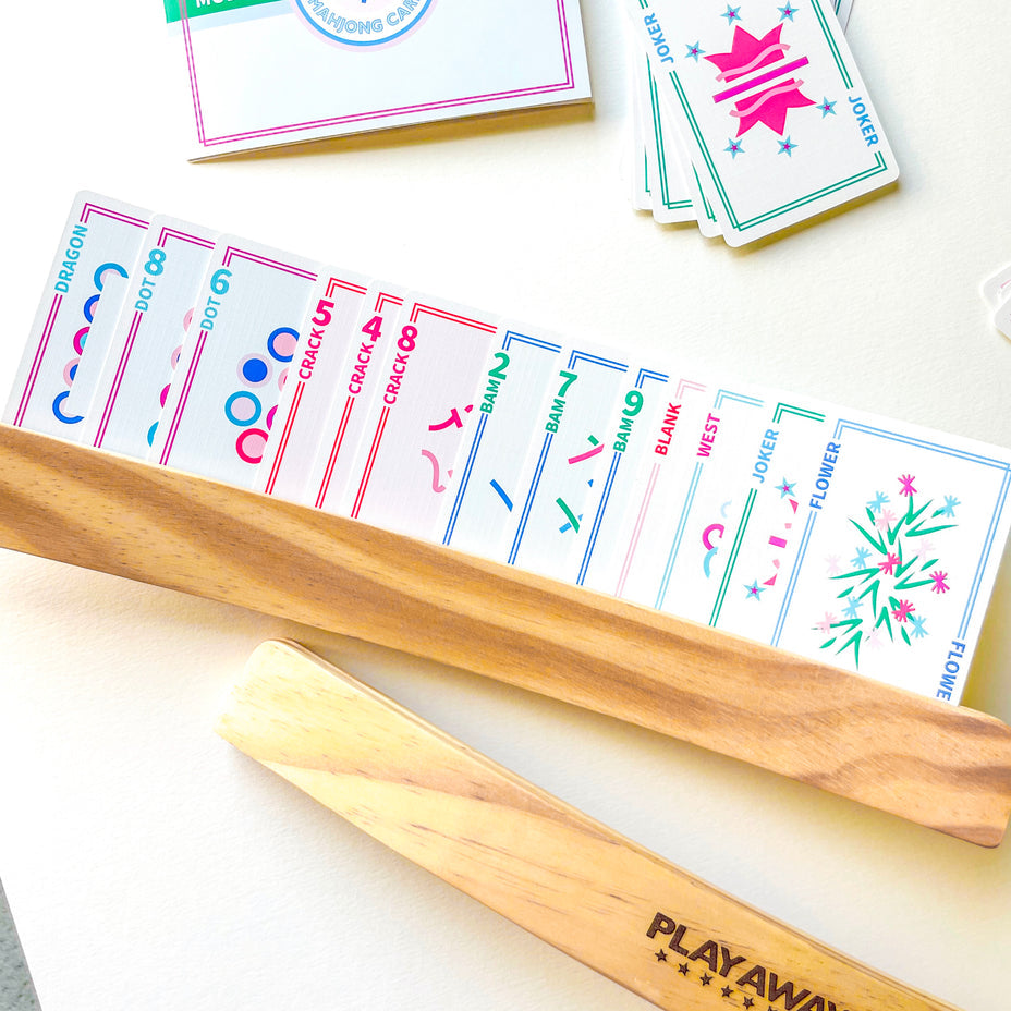 A wooden rack displays colorful Play Away Cards Mahjong Playing Cards in a row, with a "PLAYAWAY" wooden stick in front and other cards scattered on the table.