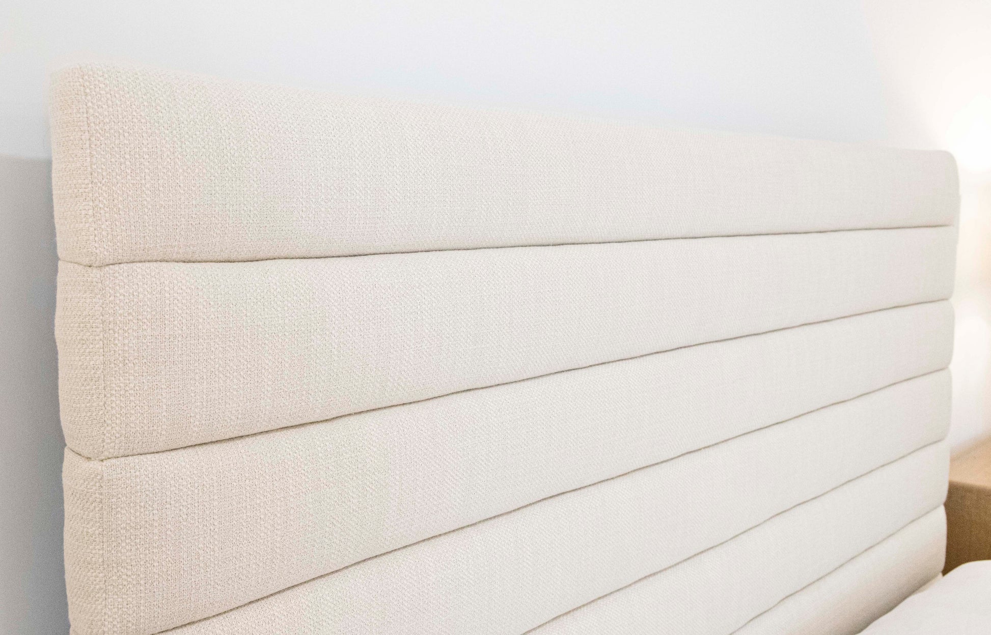 A beige, upholstered headboard with a horizontally stitched design, reminiscent of EJ Schrader's Channel Bed, King elegance, attached to a bed frame against a light-colored wall.