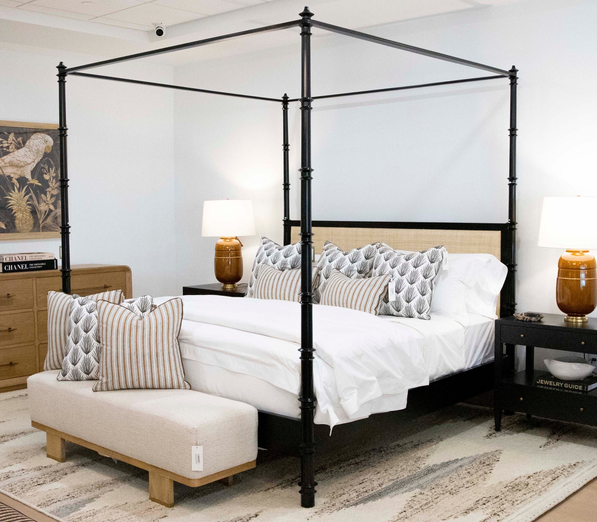 A neatly made four-poster Willa Raffia Bed, King by Oly with an upholstered headboard, white linens, and patterned throw pillows. The room includes two brown lamps on nightstands, a wooden dresser, and a small upholstered bench at the foot of the bed.