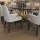 A modern Dakota Dining Table by Julian Chichester with a dark, oval top and Dante Brass base is surrounded by six white and black speckled upholstered chairs on a wooden floor. Three green decorative bowls sit on the table.