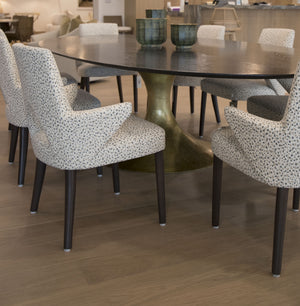 A modern Dakota Dining Table by Julian Chichester with a dark, oval top and Dante Brass base is surrounded by six white and black speckled upholstered chairs on a wooden floor. Three green decorative bowls sit on the table.