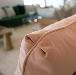 Close-up of a peach-colored Hyeres Peche Pillow, custom down-filled with a wrinkled edge, set against a blurred background of a living room with other furniture. This 22" x 22" cushion by Associated Design adds a cozy touch to the space.
