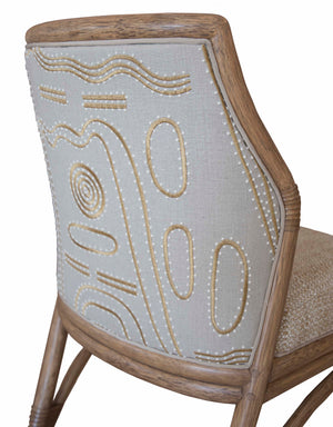 Close-up view of a McGuire Lantana Side Chair, Mushroom with intricate embroidery on the backrest, featuring abstract patterns in cream and beige tones, set within a light wooden frame. The furniture dimensions are 21.25"W and 23"D.