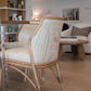 A McGuire Lantana Side Chair, Mushroom, measuring 21.25"W x 23"D, stands next to a glass table in a modern living room. Shelves with various items and a round wooden coffee table with books are visible in the background.