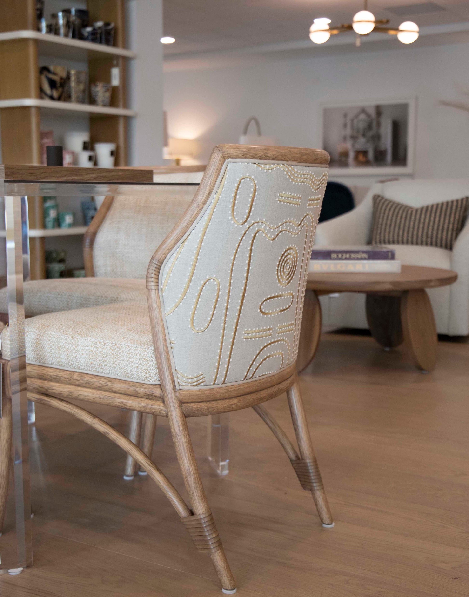 A McGuire Lantana Side Chair, Mushroom, measuring 21.25"W x 23"D, stands next to a glass table in a modern living room. Shelves with various items and a round wooden coffee table with books are visible in the background.