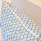 A close-up of the 22" x 22" Burma Sea Glass Outdoor Pillow by Hive Custom Pillows, featuring a blue hue with a white leaf pattern, resting on a light-colored chair.