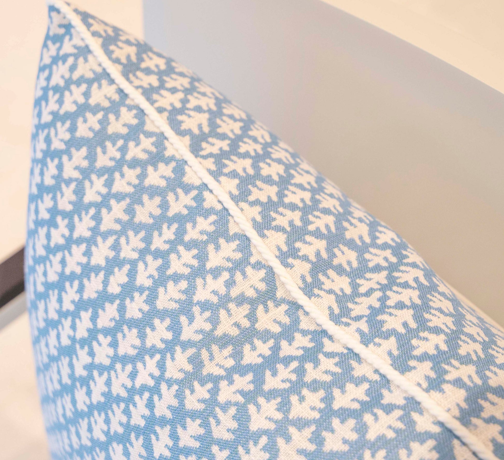A close-up of the 22" x 22" Burma Sea Glass Outdoor Pillow by Hive Custom Pillows, featuring a blue hue with a white leaf pattern, resting on a light-colored chair.