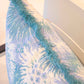 Close-up of the Hive Custom Pillows Bimini Sea Glass Outdoor Pillow, 22x22, featuring a blue and white floral pattern with textured blue fringe, displayed on a light-colored chair.