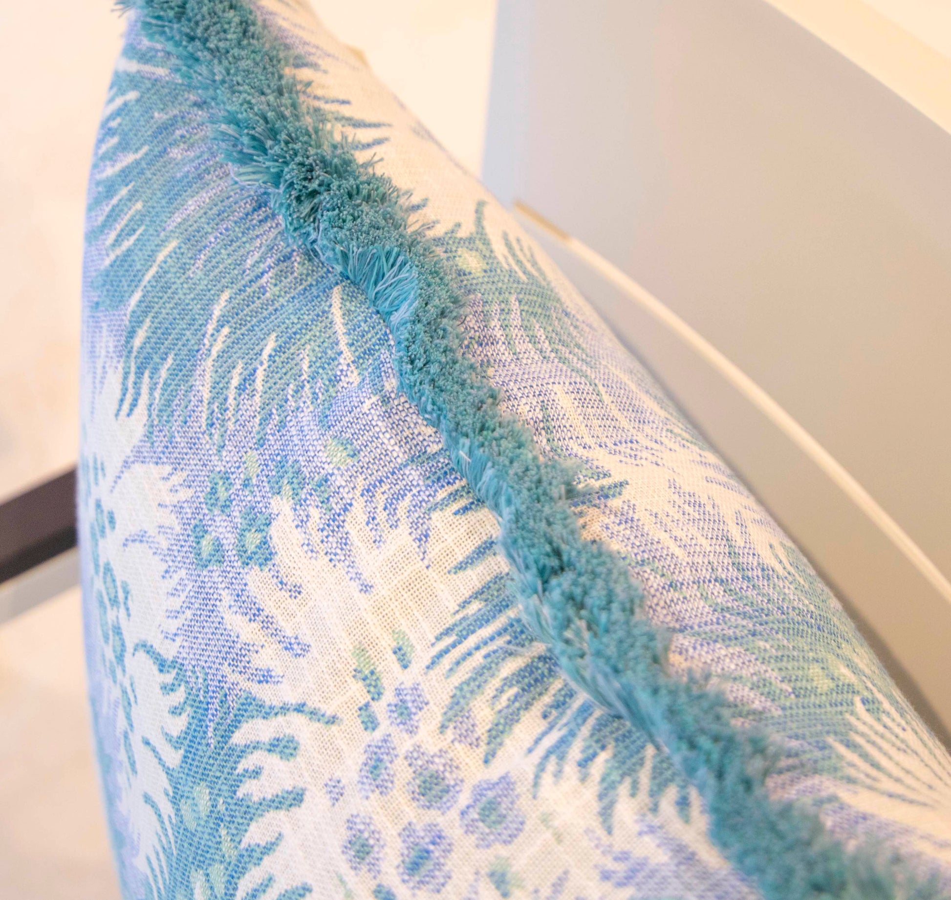 Close-up of the Hive Custom Pillows Bimini Sea Glass Outdoor Pillow, 22x22, featuring a blue and white floral pattern with textured blue fringe, displayed on a light-colored chair.