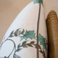 A close-up of the Poppy Stripe Aqua Lumbar Pillow by Hive Custom Pillows, featuring green and gray botanical designs, resting against a wicker chair's curved arm.