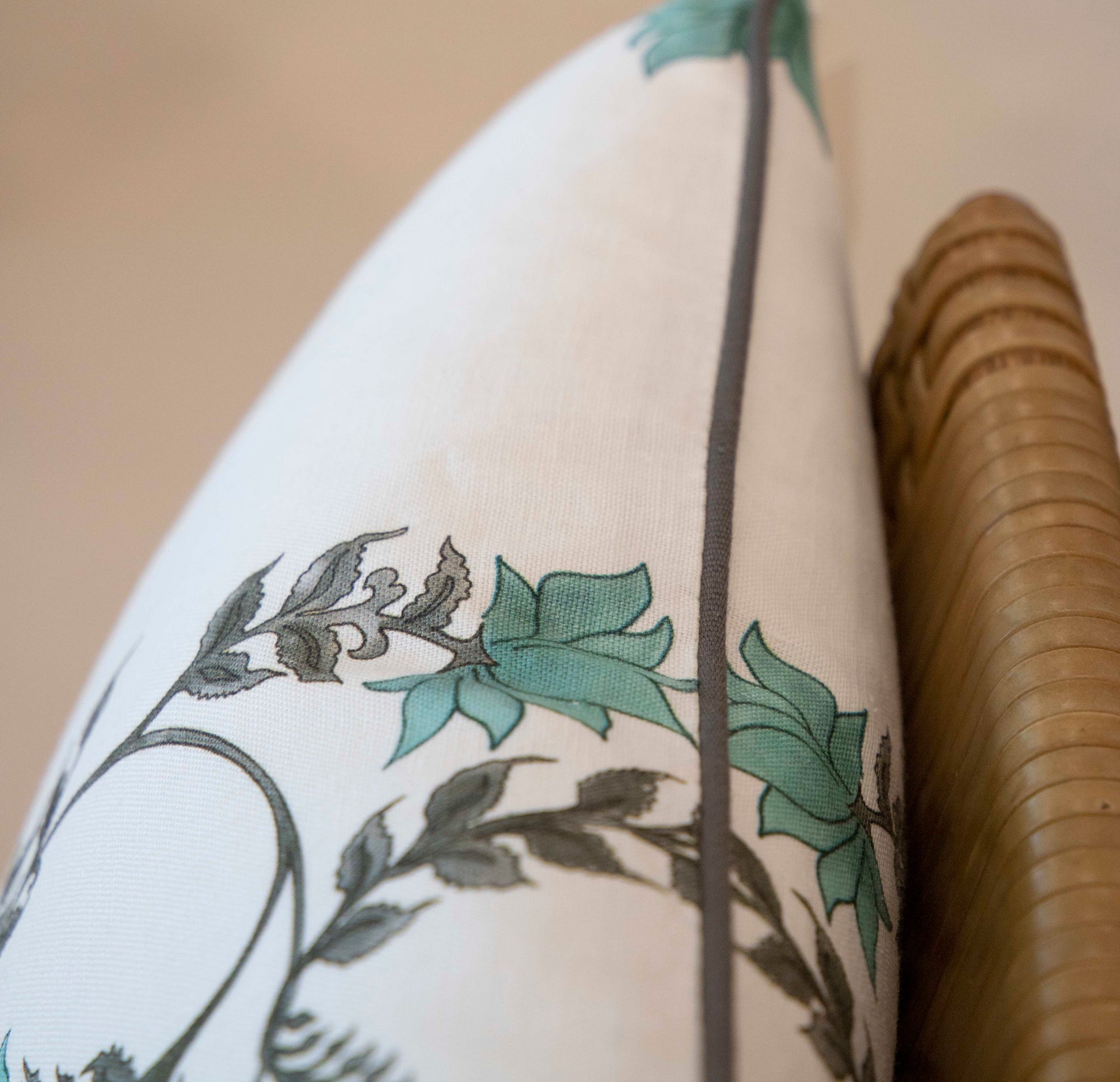 A close-up of the Poppy Stripe Aqua Lumbar Pillow by Hive Custom Pillows, featuring green and gray botanical designs, resting against a wicker chair's curved arm.