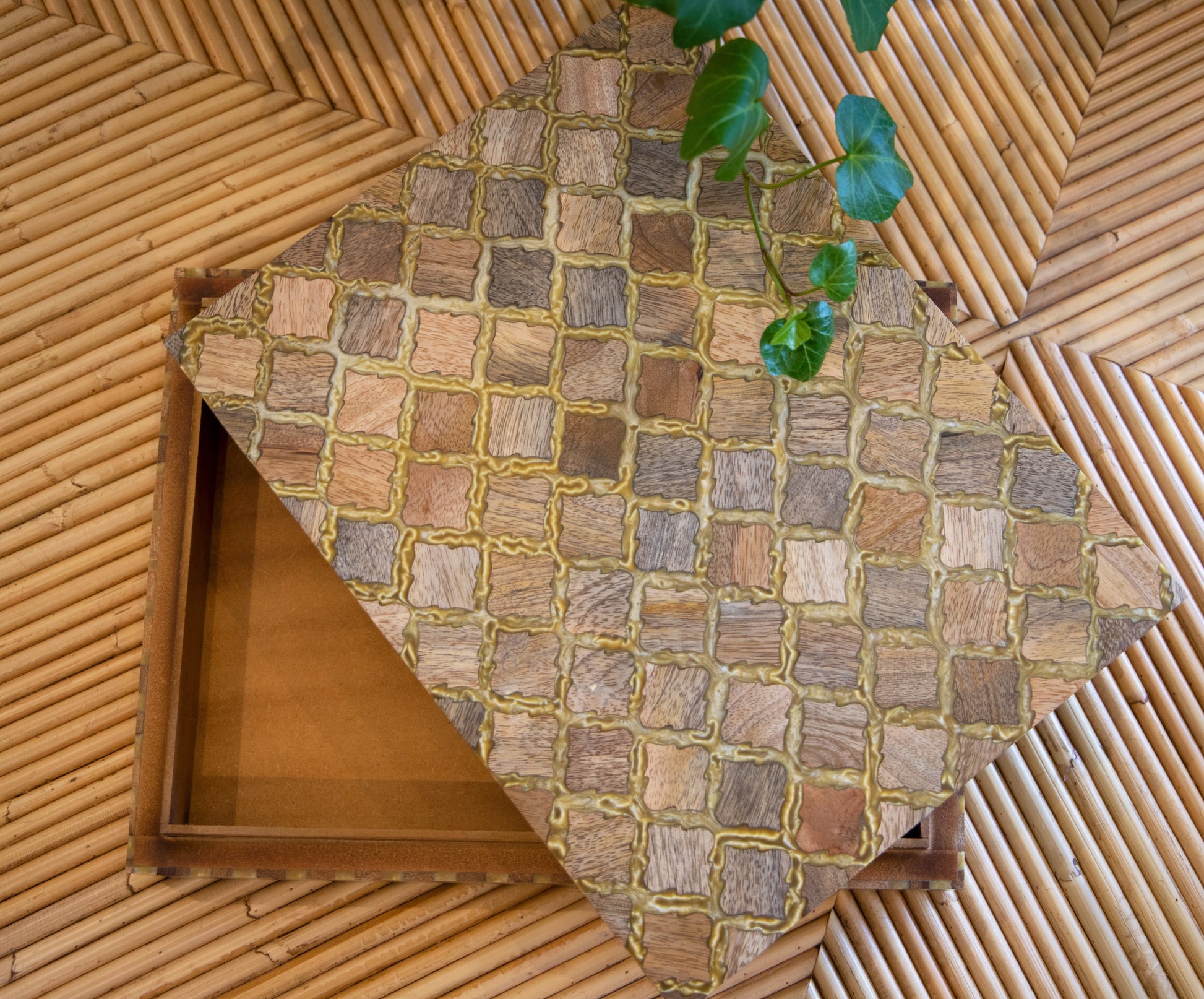 A decorative wooden box, the Bojay Diamond Box in Medium, featuring a diamond pattern and mother of pearl inlay is partially open on a bamboo mat, with a green plant extending into the frame.