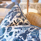Close-up of the Damour Blues Pillow by Hive Custom Pillows, a handmade, down-filled cushion featuring a blue and white floral pattern, displayed on a woven chair. Crafted in the USA.