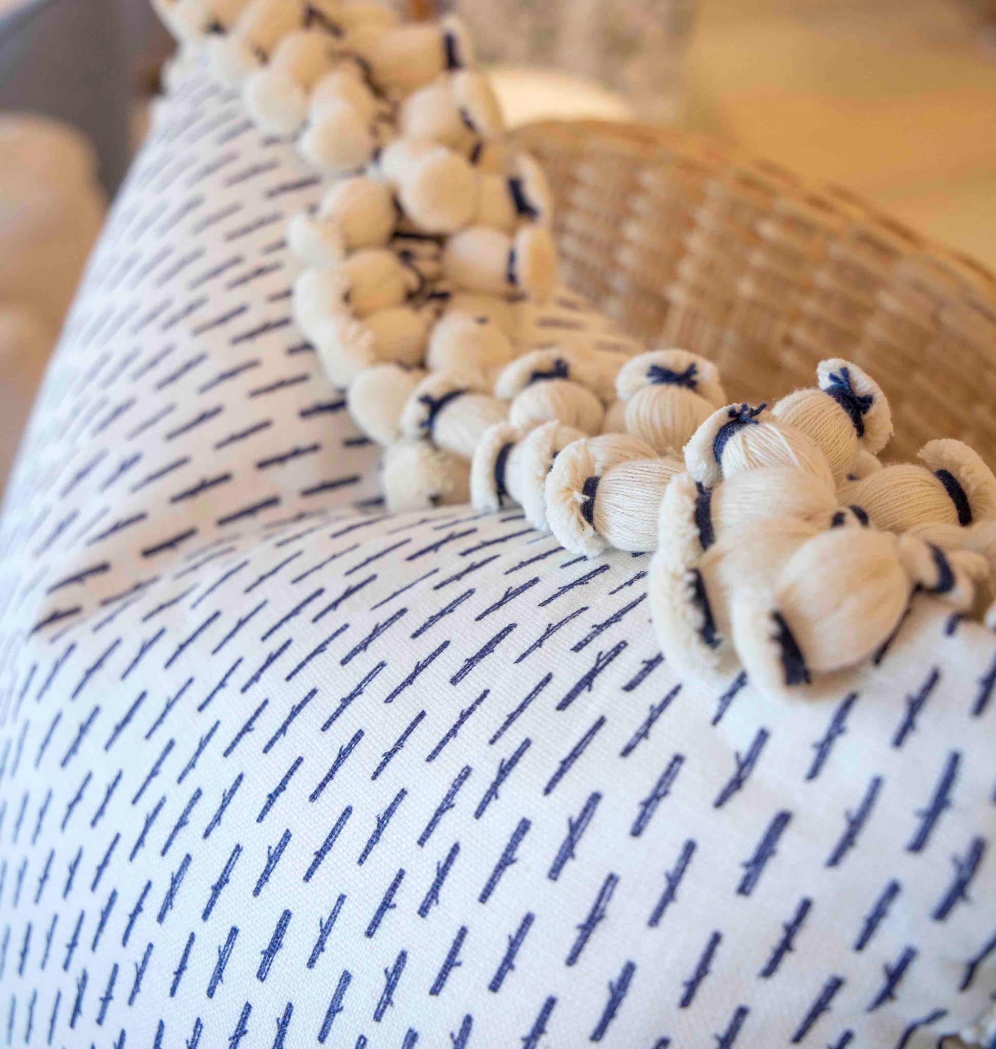 Close-up of the Little Bamboo Navy Pillow by Hive Custom Pillows, a handmade down-filled pillow with a blue dashed pattern and cream pom-pom trim, resting next to a woven basket.