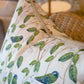 Close-up of the Hummingbirds White Pillow by Associated Design, featuring green leaves and blue-green birds, placed beside a woven chair with a softly blurred lamp in the background.