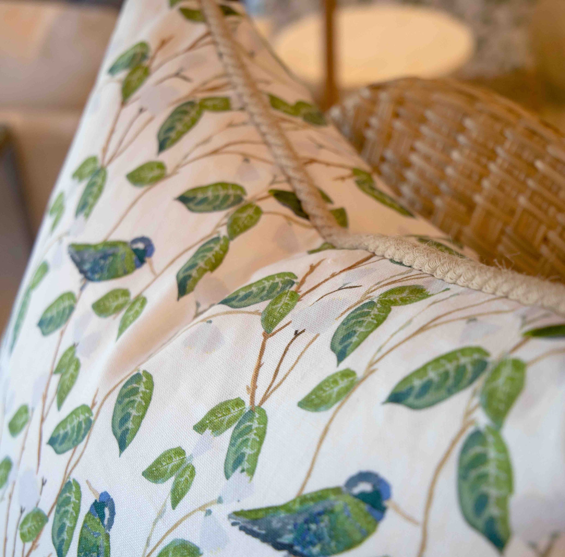 Close-up of the Hummingbirds White Pillow by Associated Design, featuring green leaves and blue-green birds, placed beside a woven chair with a softly blurred lamp in the background.