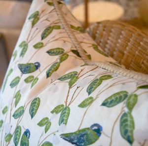Close-up of the Hummingbirds White Pillow by Associated Design, handmade in the USA with a green leaf and blue bird pattern, shown on a wicker chair with a lamp softly blurred in the background.