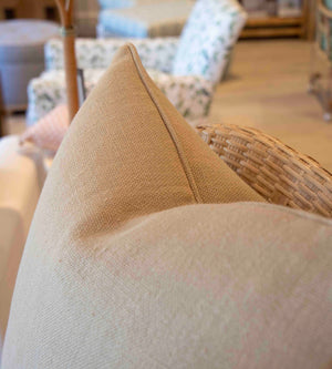 Close-up of a Cobble Farthing Pillow by Associated Design, featuring a handmade beige texture and down filling, resting on a wicker chair with blurred chairs and wooden floor in the background.