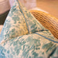 A close-up of a handmade wicker chair features the Heather Moss Pillow from Beleo Interiors LLC, with a decorative down-filled cushion showcasing a textured blue and white pattern, adding an artisanal touch to the scene.