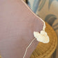 A Royal Bohemian Heather Pillow by Associated Design, custom-made in purple with white piping and tassels, is seen on a wicker chair with plush down filling, set against leaf-patterned wallpaper.