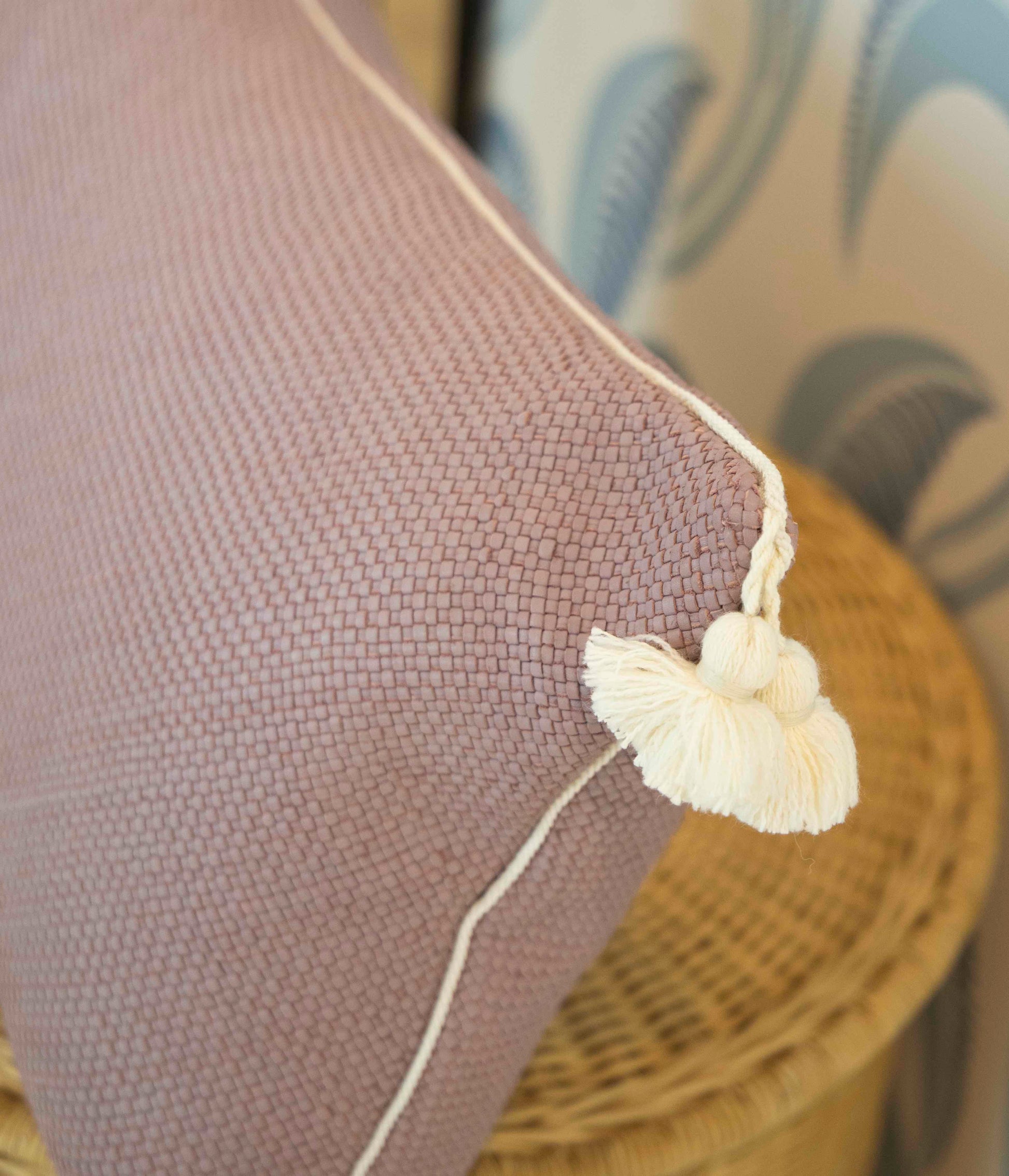 A Royal Bohemian Heather Pillow by Associated Design, custom-made in purple with white piping and tassels, is seen on a wicker chair with plush down filling, set against leaf-patterned wallpaper.
