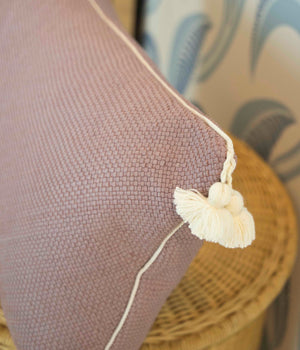 A Royal Bohemian Heather Pillow by Associated Design, custom-made in purple with white piping and tassels, is seen on a wicker chair with plush down filling, set against leaf-patterned wallpaper.