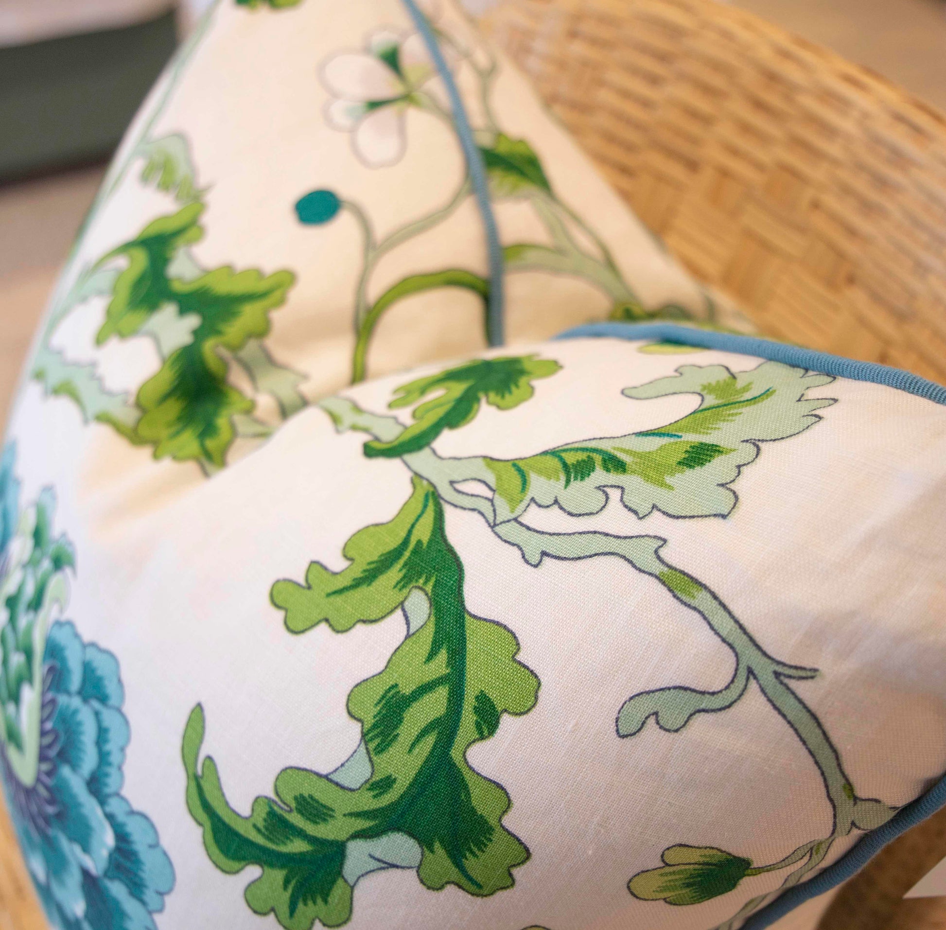 Close-up of two New Athos Spring Pillows by Hive Custom Pillows, featuring green and blue botanical patterns on a cream background, placed on a woven chair. Expertly crafted for comfort and style.