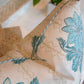 Close-up of the Lucknow Seafoam Pillow by Hive Custom Pillows, a handmade cream-colored cushion with blue floral patterns and piping, shown on a textured surface with green leaves in the background.