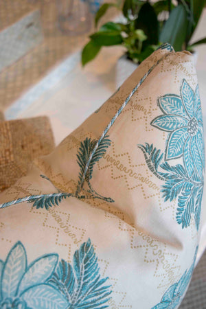 Close-up of the Lucknow Seafoam Pillow by Hive Custom Pillows, a handmade cream-colored cushion with blue floral patterns and piping, shown on a textured surface with green leaves in the background.