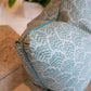 A close-up of the Jhinuk Teal Pillow by Hive Custom Pillows, a 22" x 22" blue and white patterned throw pillow with blue piping, handmade in the USA and down-filled, placed on a beige couch with a potted plant in the background.