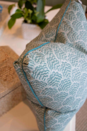 A close-up of the Jhinuk Teal Pillow by Hive Custom Pillows, a 22" x 22" blue and white patterned throw pillow with blue piping, handmade in the USA and down-filled, placed on a beige couch with a potted plant in the background.