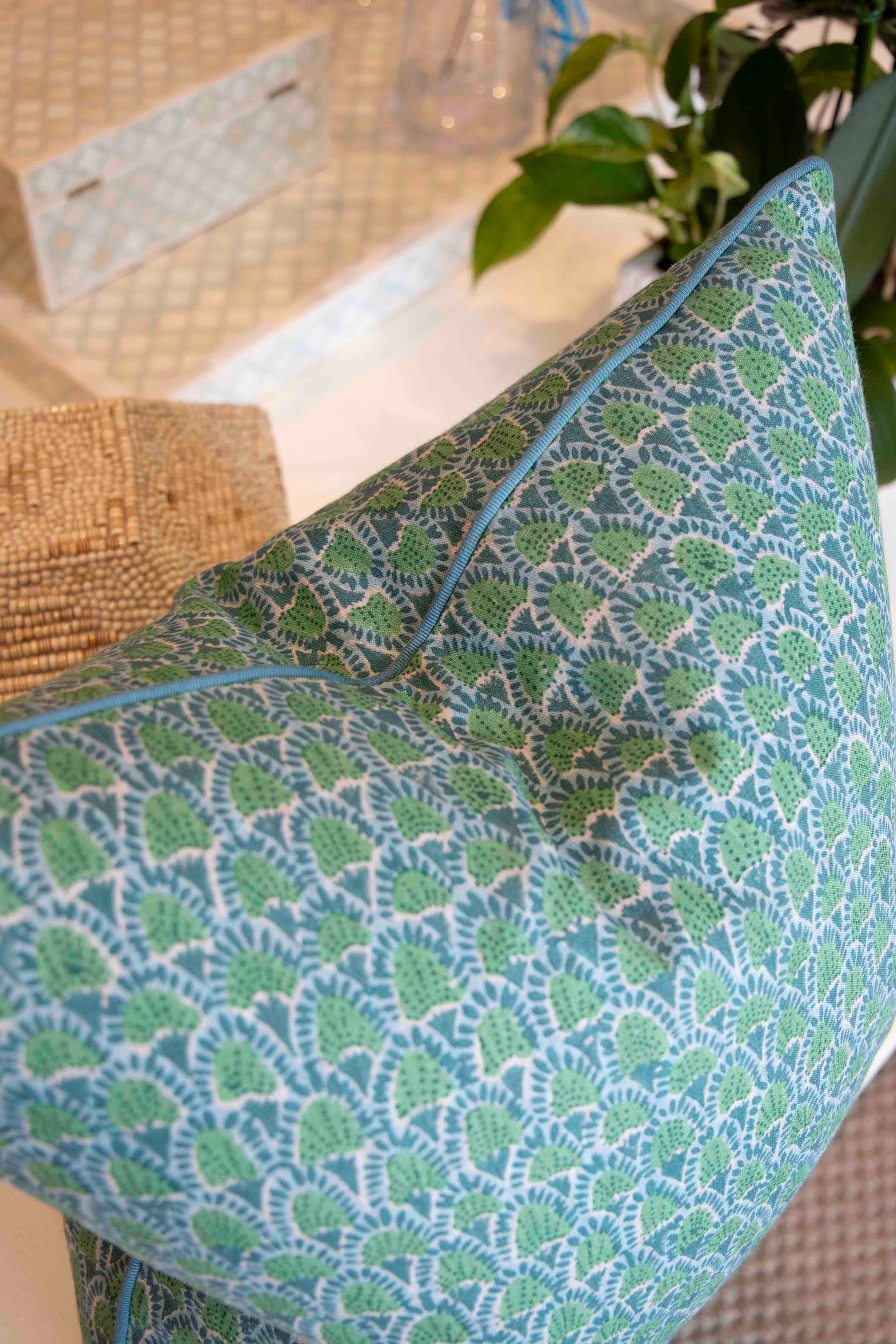 The Scopello Emerald Linen Pillow by Hive Custom Pillows, handmade in the USA with down filling and a blue-green leaf pattern, rests on a light surface beside a plant and woven decor.