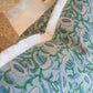 Close-up of the Marbella Linen Emerald Pillow by Hive Custom Pillows, featuring a blue and green floral design with white fringe trim, displayed on furniture with a plant in the background.