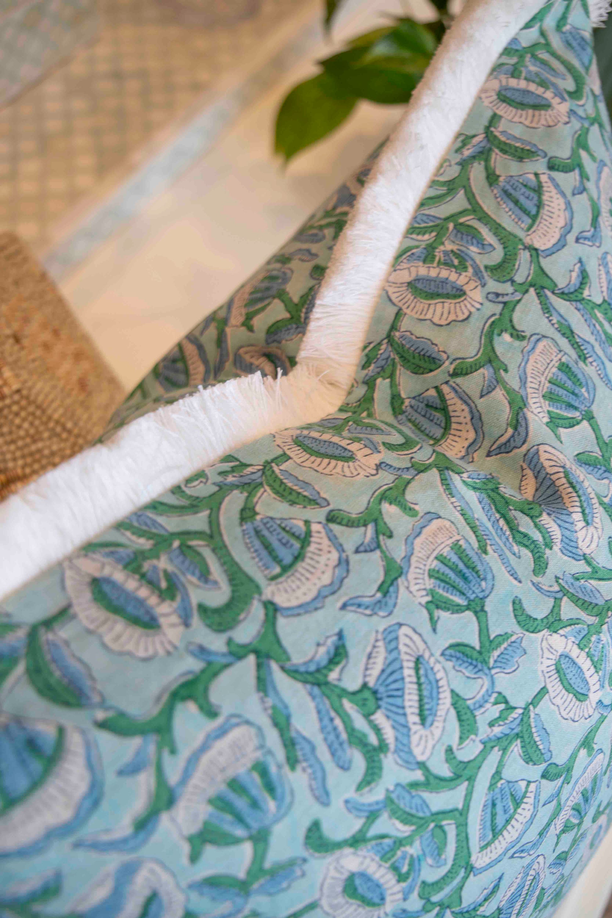 Close-up of the Marbella Linen Emerald Pillow by Hive Custom Pillows, featuring a blue and green floral design with white fringe trim, displayed on furniture with a plant in the background.