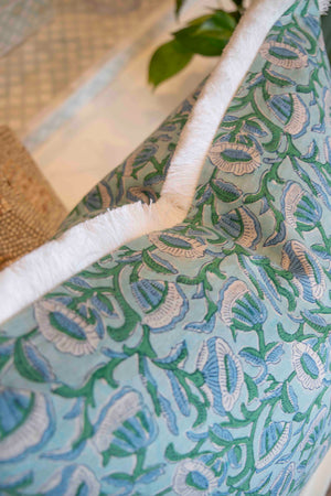Close-up of the Marbella Linen Emerald Pillow by Hive Custom Pillows, featuring a blue and green floral design with white fringe trim, displayed on furniture with a plant in the background.