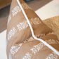 The Petite Frond Camel Pillow by Hive Custom Pillows is handmade in the USA with white leaf patterns and white piping on brown fabric, shown here on a light-colored surface.