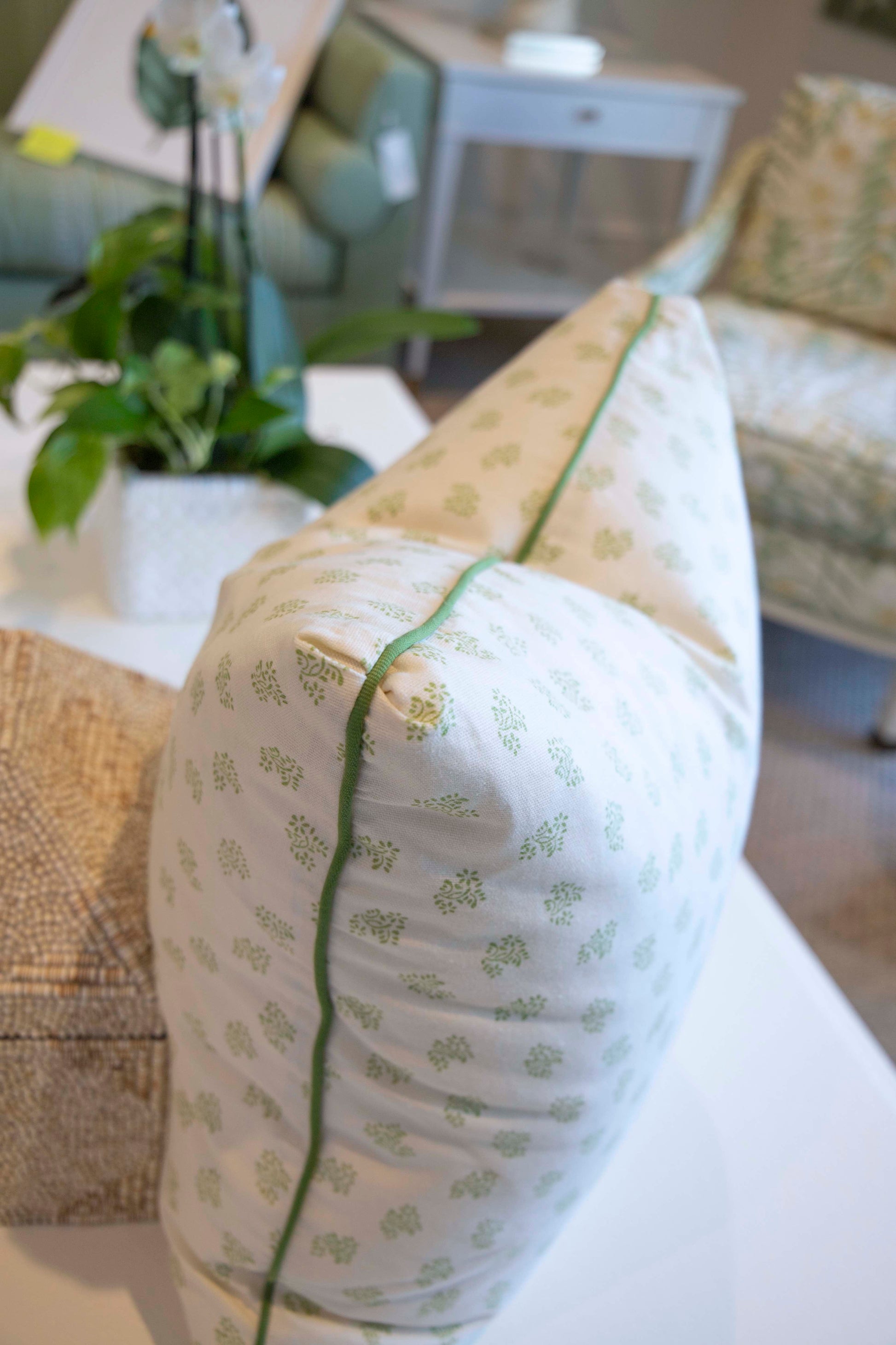 A close-up of the Dolly Flower Lettuce Green Pillow by Associated Design, featuring a light pattern and green piping, rests on a white table in a furnished living room. Handmade in the USA for an elegant touch.