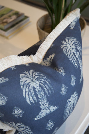The Mandalay Indigo Pillow by Hive Custom Pillows, handmade in the USA, features blue down-filled fabric with white palm tree patterns and beige fringe, shown on a white surface with a plant and books in the background.