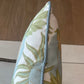 A close-up of the Carnival Celeste Outdoor Pillow by Associated Design, featuring a blue and green leaf pattern with blue braided edging, placed on a chair with tile and wood flooring in the background.