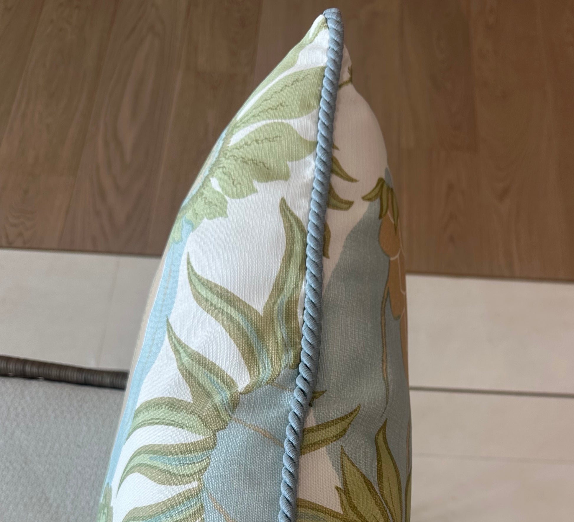 A close-up of the Carnival Celeste Outdoor Pillow by Associated Design, featuring a blue and green leaf pattern with blue braided edging, placed on a chair with tile and wood flooring in the background.
