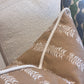 The Petite Frond Pillow by Hive Custom Pillows features a tan cover with a white leaf design and rope trim, crafted from fine materials. Shown in close-up, it rests on a textured light sofa beside another patterned pillow.