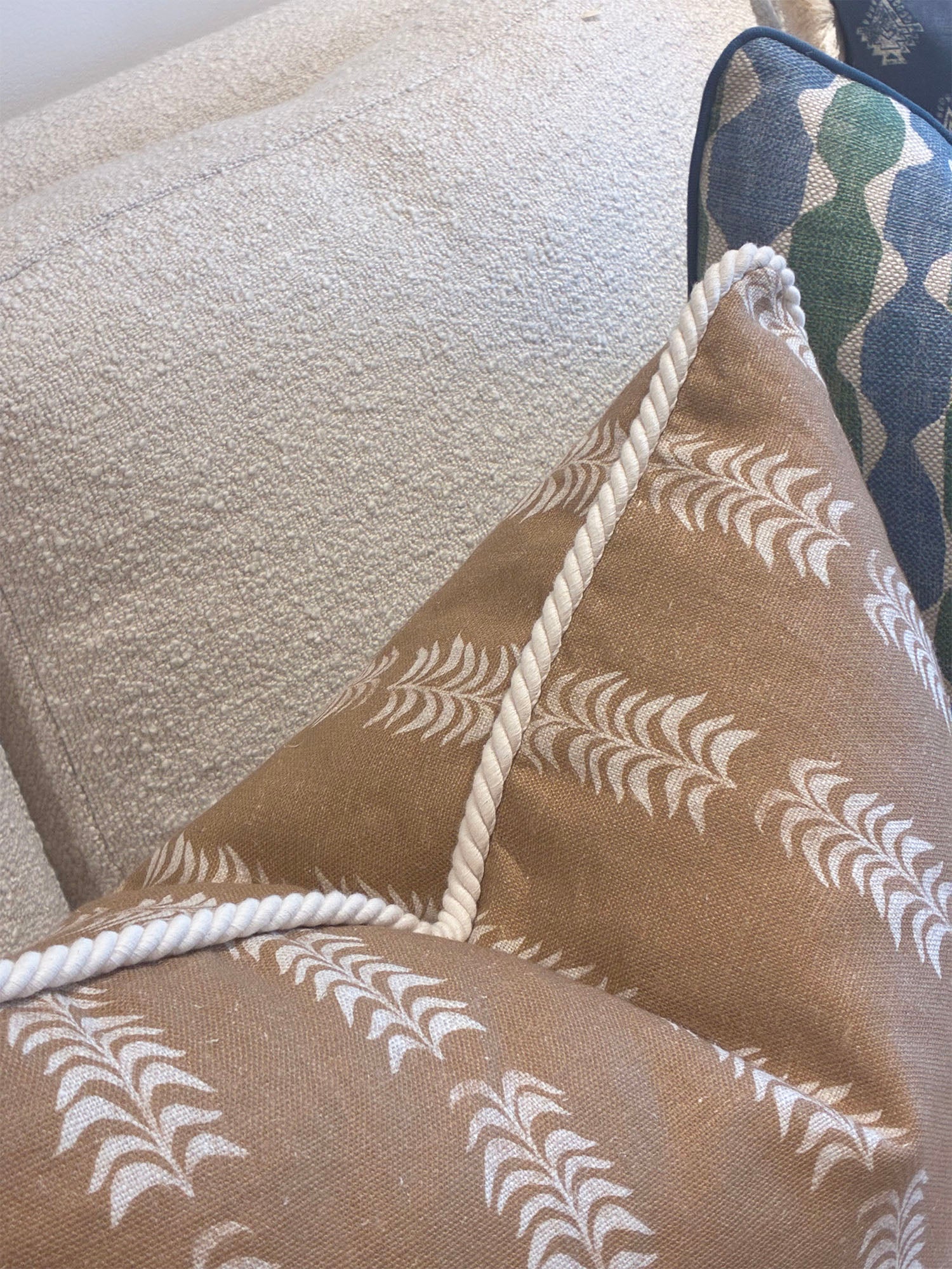 The Petite Frond Pillow by Hive Custom Pillows features a tan cover with a white leaf design and rope trim, crafted from fine materials. Shown in close-up, it rests on a textured light sofa beside another patterned pillow.